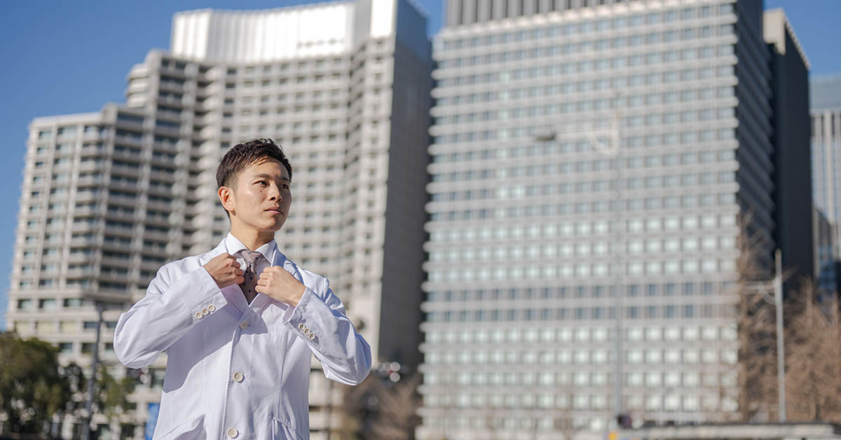 製薬会社を背景に立つ白衣を着た薬剤師の男性