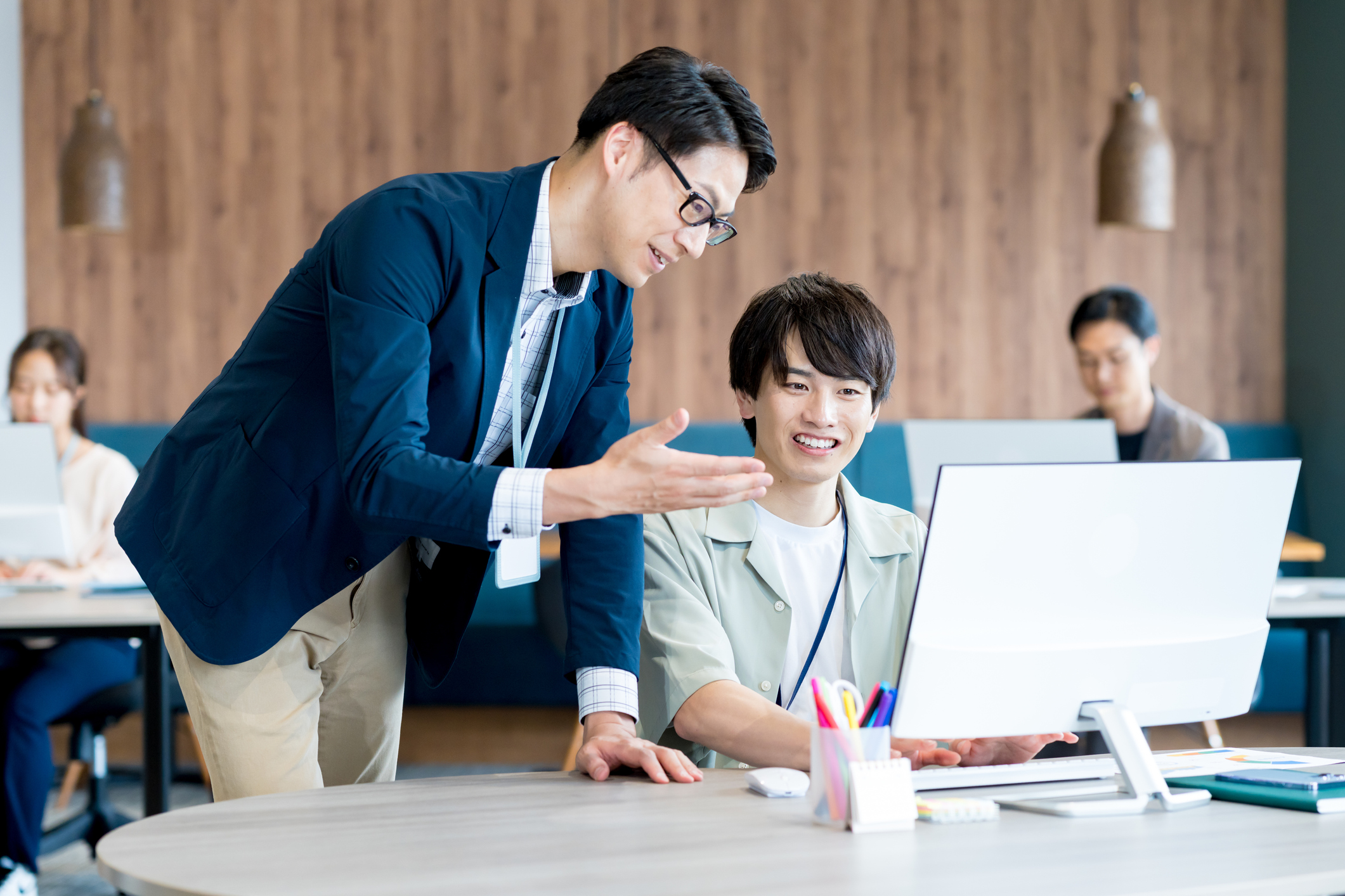 スタートアップオフィスで若手社員とベテラン社員が並んでパソコン画面を見ながら共同作業をしている様子。