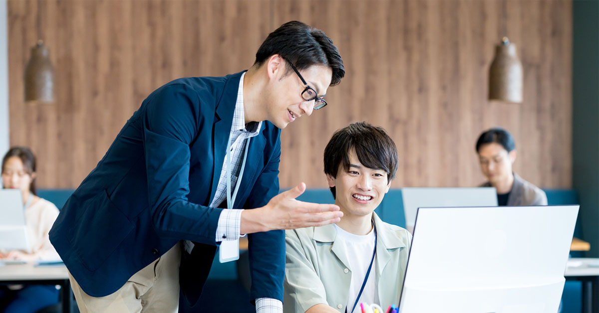 スタートアップオフィスで若手社員とベテラン社員が並んでパソコン画面を見ながら共同作業をしている様子。